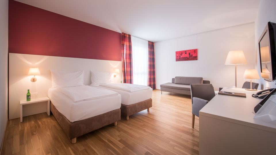Zwei Betten stehen auf hellem Holzboden vor rotweisser Wand.