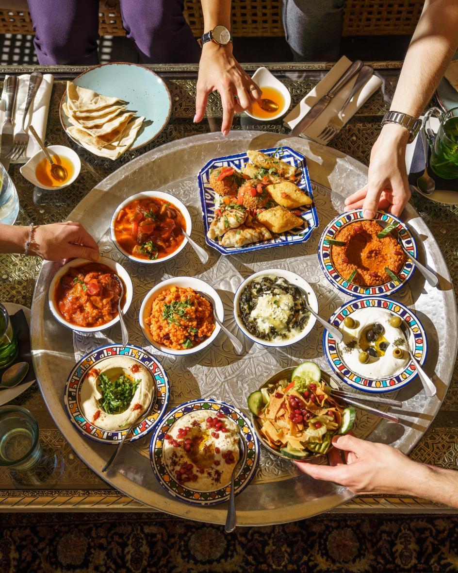 Mezzeplatte steht in der Mitte eines Tisches und vier Menschen bedienen sich an den bunt angerichteten Speisen.