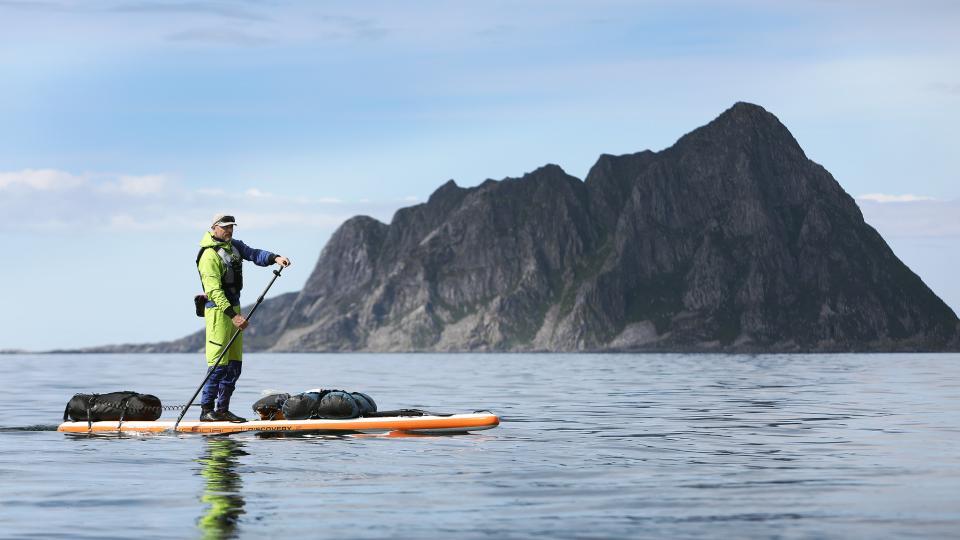 Ein Stand-up-Paddler ist mit Gepäck auf dem Meer unterwegs. Im Hintergrund ist eine Felsinsel zu sehen.