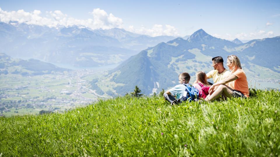 Eine Familie mit zwei Kindern sitzt im Gras und Blick in die Urner Bergwelt. Im Hintergrund ist der Vierwaldstättersee und die Rigi zu sehen.