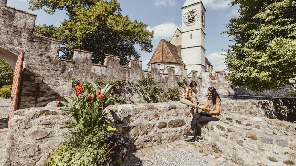 Zwei Frauen sitzen auf einer Steinmauer und studieren eine Broschüre. Im Hintergrund ist eine Kirche zu sehen.