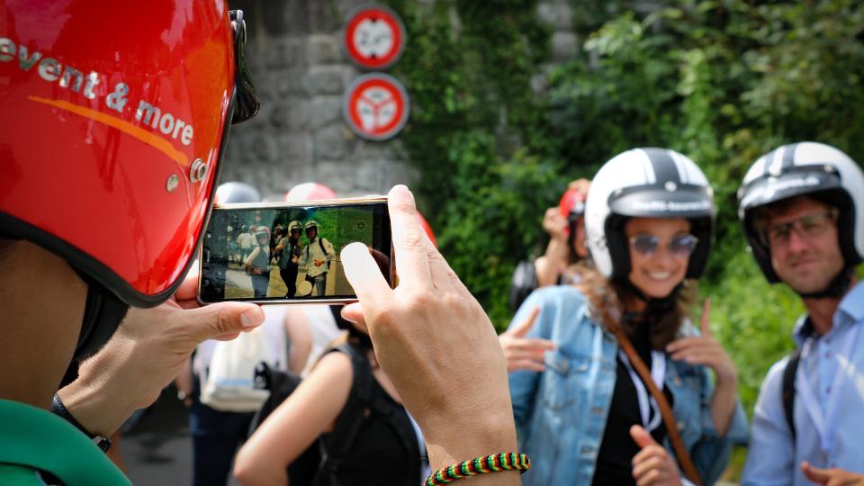 Ein Paar mit Motorradhelmen posiert vor grünen Büschen und wir mit einem Smartphone fotografiert.