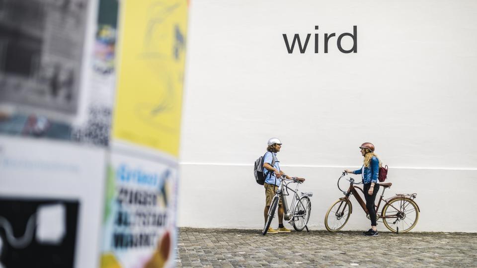 Zwei Radfahrende stehen vor einer wissen Wand mit dem Schriftzug wird des Kunsthauses Zug.