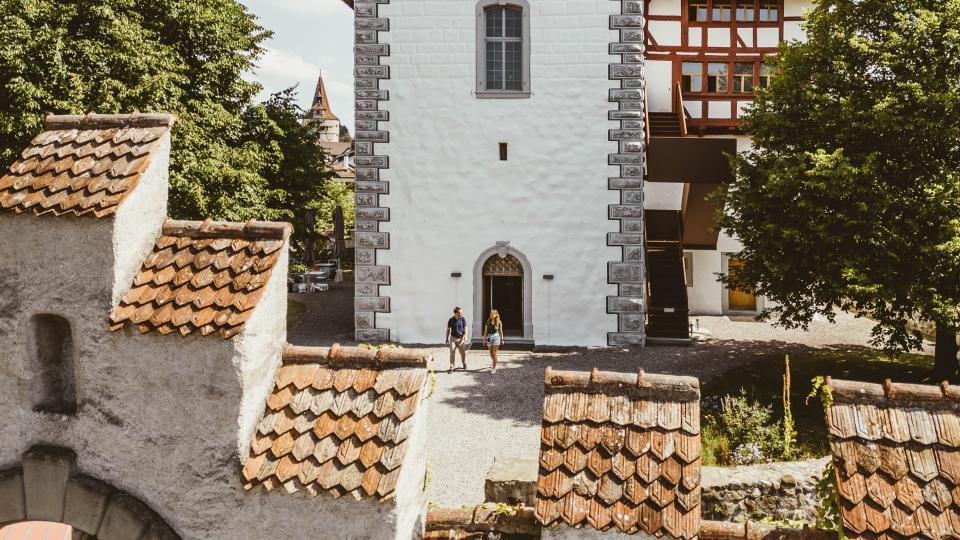 Ein Mann und eine Frau laufen aus dem historischen Steinturm des Museum Burg Zug auf dem Kiesweg Richtung Torbogen.
