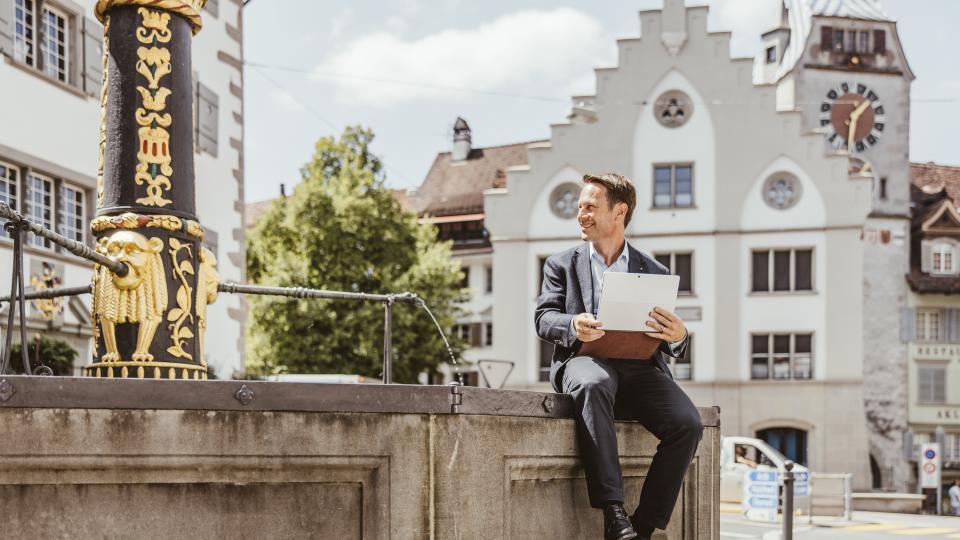 Ein Geschäftsmann sitzt auf einem Brunnen in der Altstadt Zug. Im Hintergrund ist der Zytturm zu sehen.
