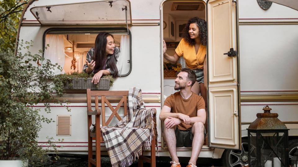 Zwei Damen und ein Herr stehen und sitzen in einem Wohnwagen.