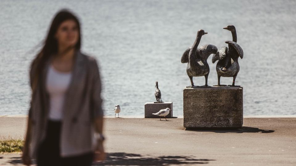 Ein Geschäftsfrau spaziert am Seeufer entlang. Im Hintergrund sind Statuen von Vögeln zu sehen.