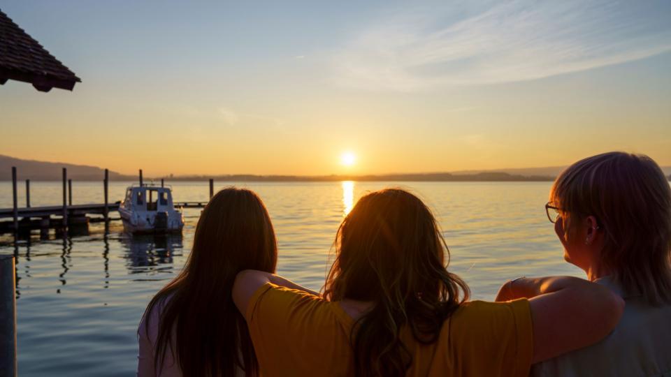Drei Frauen sitzen am Ufer des Zugersee und schauen in den Sonnenuntergang