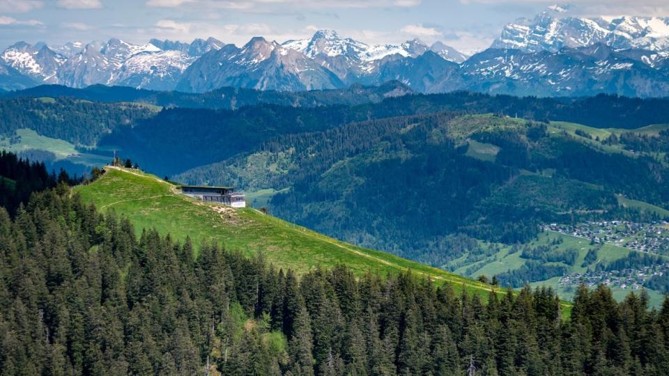 Das Bergrestaurant steht auf einer grünen Wiese und im Hintergrund sind die verschneiten Gipfel der Glarner Alpen zu sehen.