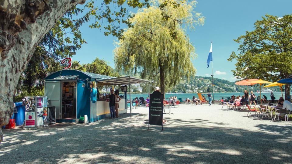 Eine Strandbuvette steht unter Bäumen am Seeufer. Menschen sitzen unter Sonnenschirmen oder entspannen im Liegestuhl.