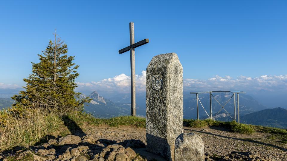 Der Grenzstein und das Gipfelkreuz thronen auf dem Wildspitz. Links ist der grosse Mythen sowie eine Wal- und Wiesenlandschaft zu sehen.