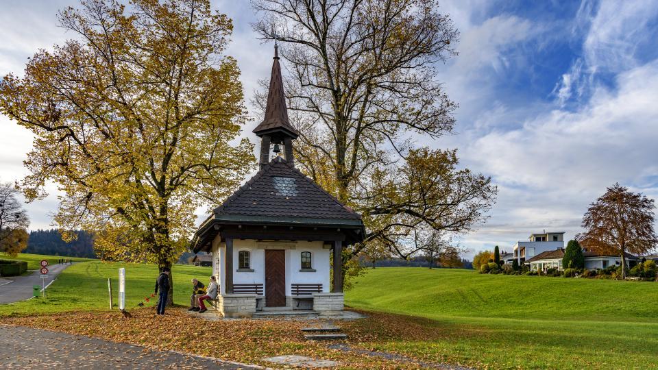 Die kleine Kapelle steht zwischen zwei herbstlichen Bäumen auf einer grünen Wiese. Auf der Bank vor der Kapelle sitzen zwei Menschen und unterhalten sich mit einem Spazierenden.