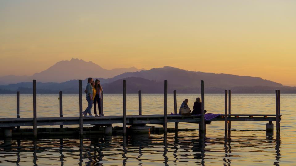 Drei Frauen stehen auf einem Steg über dem See und geniessen die Abendstimmung. Im Hintergrund leuchtet der Pilatus im Abendrot.