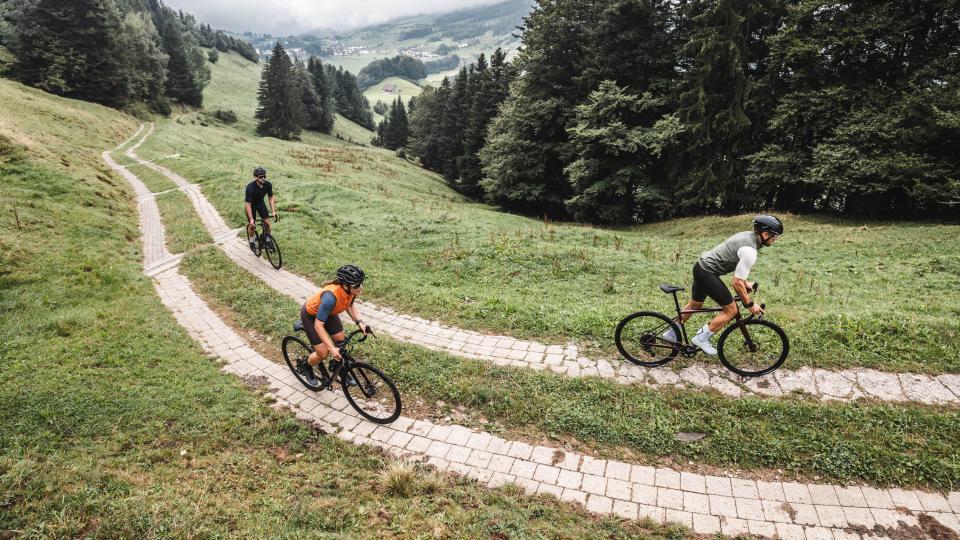 Eine Gruppe Gravelfahrende pedalieren auf einer Kopfsteinstrasse durch grüne Alpwiesen den Berg hoch.