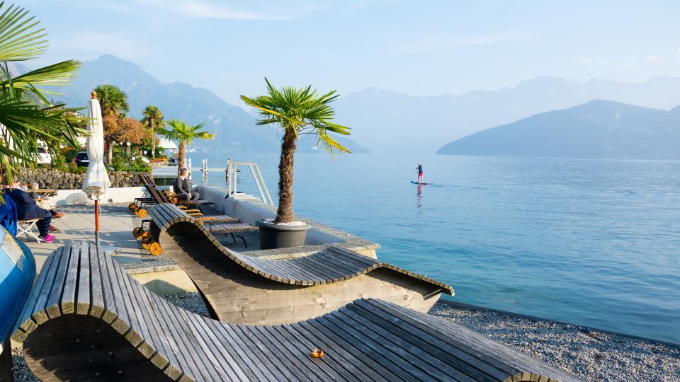 Breite Holzliegestühle stehen am Ufer des See. Weisse Sonnenschirme und Palmen sorgen für eine mediterrane Atmosphäre. 
