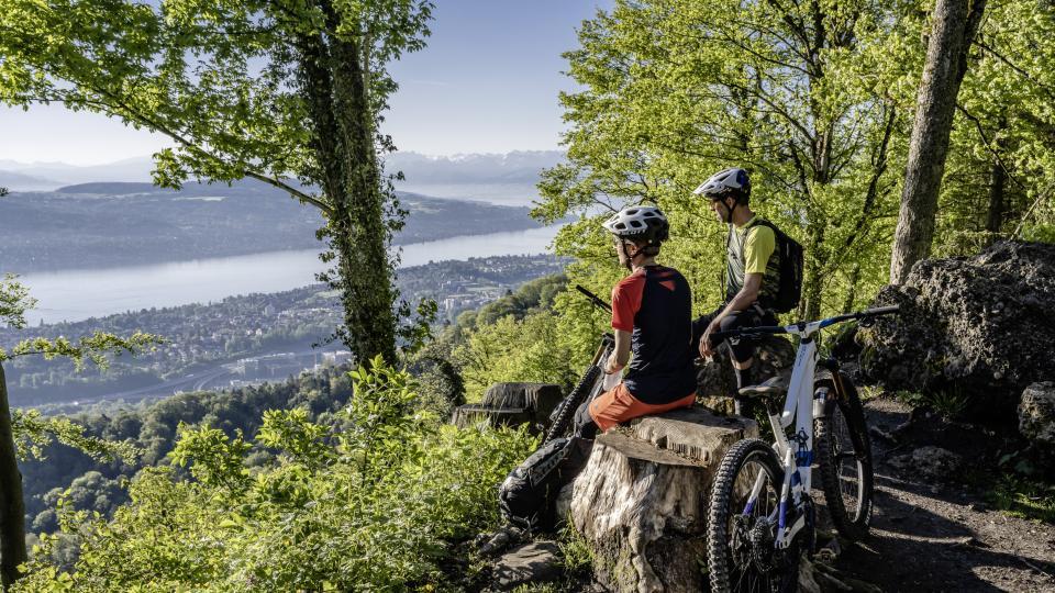 Ein mountainbike Paar sitzt auf einem Baumstrunk in einer Lichtung und Blickt auf den Zürichsee.