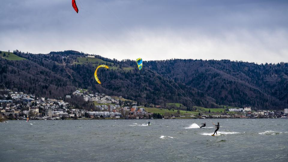 Drei Kitesurfer kiten auf dem Zugersee. Im Hintergrund ist die Zuger Altstadt sowie der Zugerberg zu sehen.