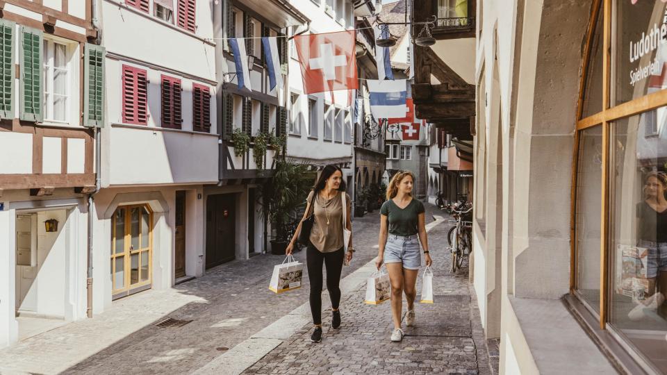 Zwei Frauen bummeln mit Einkaufstaschen auf Kofpsteinpflaster zur die Zuger Altstadt.