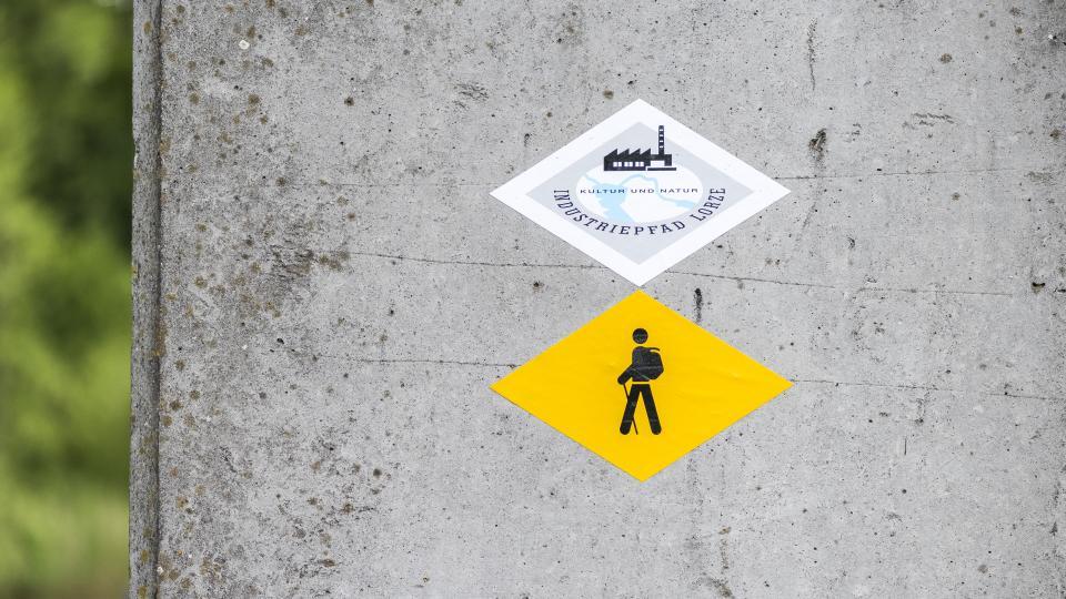 Ein Wegweiser des Industriepfads und ein Schild der Wanderwege sind an einer Betonsäule befestigt.