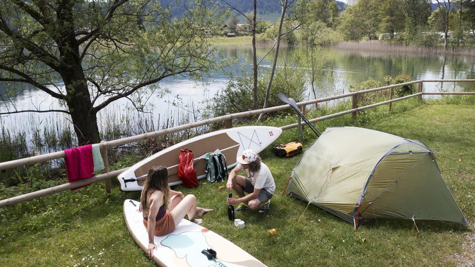 Zwei SUP-Tourende Campen am Ufer des Ägerisees. Die beiden sitzen vor dem Zelt und öffnen eine Flasche Wein.