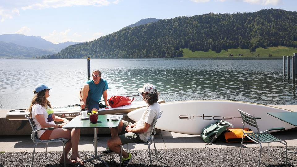 Drei Menschen sitzen an einem Tisch am See, trinken Kaffee und blicken auf die grüne Hügelandschaft.