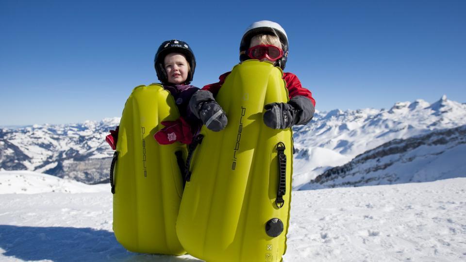 Zwei Kinder stehen in der verschneiten Winterlandschaft hinter dem Body-Board.