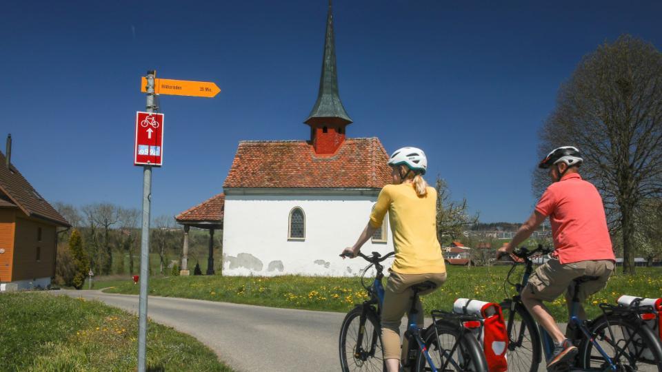 Zwei Velofahrende sind auf einer Asphaltstrasse vor einer kleinen Kapelle am Velofahren. Die Beiden sind auf der Herzroute von Zug nach Willisau unterwegs.
