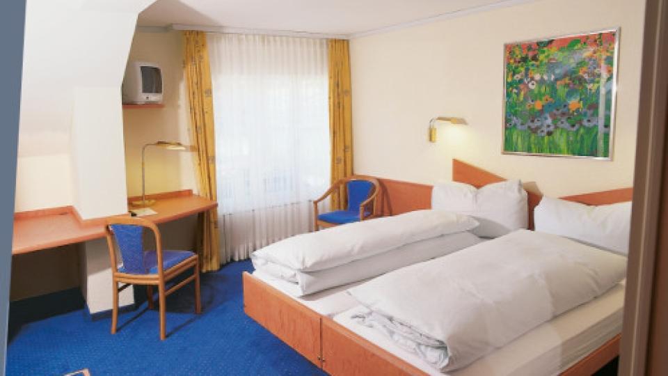 Ein Doppelbett mit weisser Bettwäsche steht vor einer weissen Wand mit einem Blumenbild. Am Boden ist blauer Teppich verlegt.