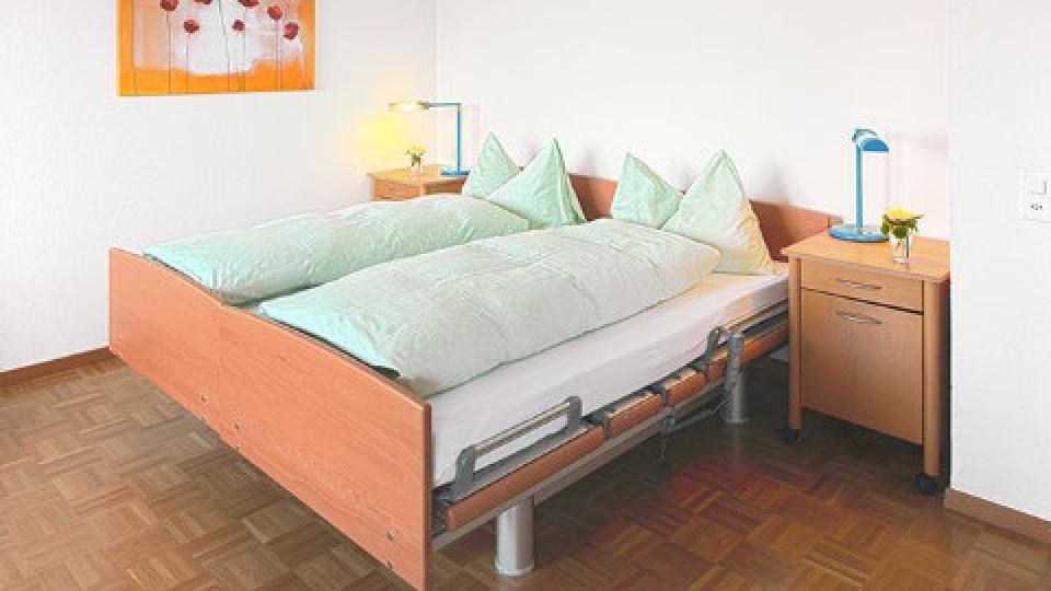 Bett mit grüner Bettwäsche steht auf Holzboden und vor weissen Wänden.