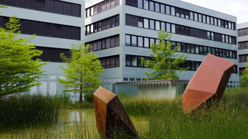 Ein Kunstobjekt aus rostigem Metall steht in einem Biotop vor Laubbäumen in einem Innenhof von Bürogebäuden.