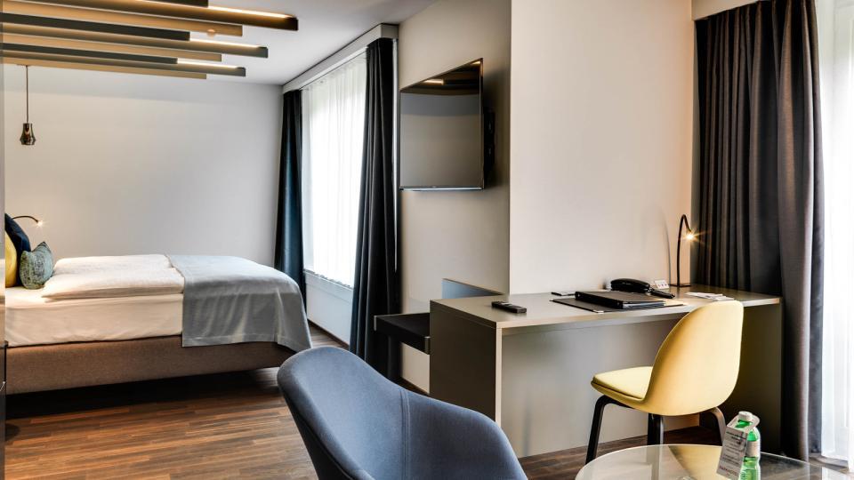 Farbig bezogenes Bett steht auf einem hellen Holzboden des Parkhotels Zug. Im Vordergrund steht das Businessabteil des Hotels.