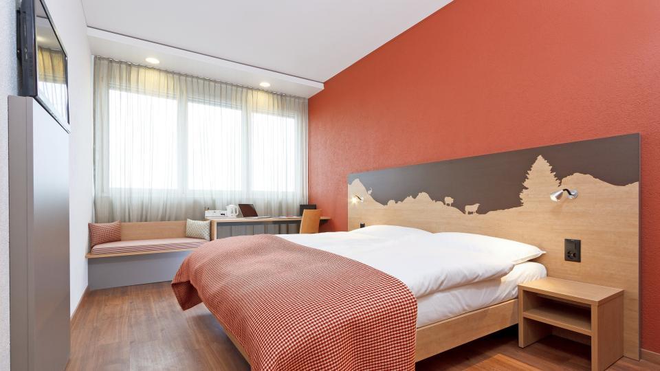 Alpenchic Doppelbett auf hellem Holzboden mit roter Wand.