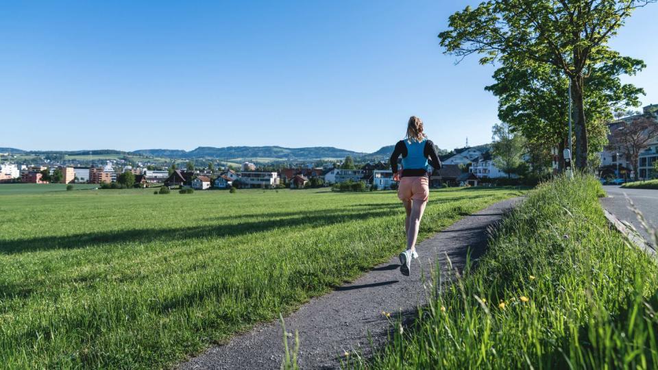Eine Frau joggt auf einem Kiesweg durch grüne Felder.