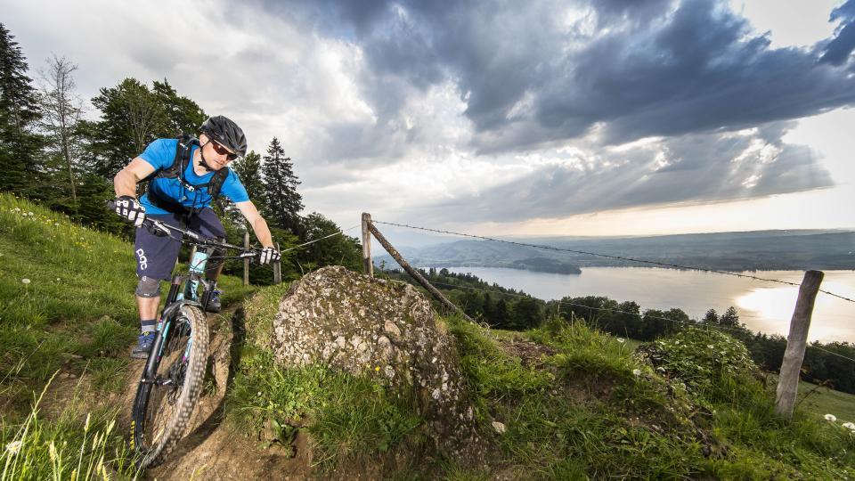 Ein Mountainbiker ist auf einer gebauten Mountainbikepiste unterwegs Im Hintergrund ist der Zugersee zu sehen.