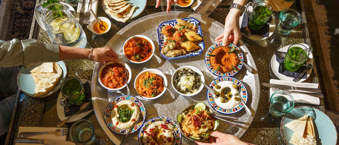Mezzeplatte steht in der Mitte eines Tisches und vier Menschen bedienen sich an den bunt angerichteten Speisen.