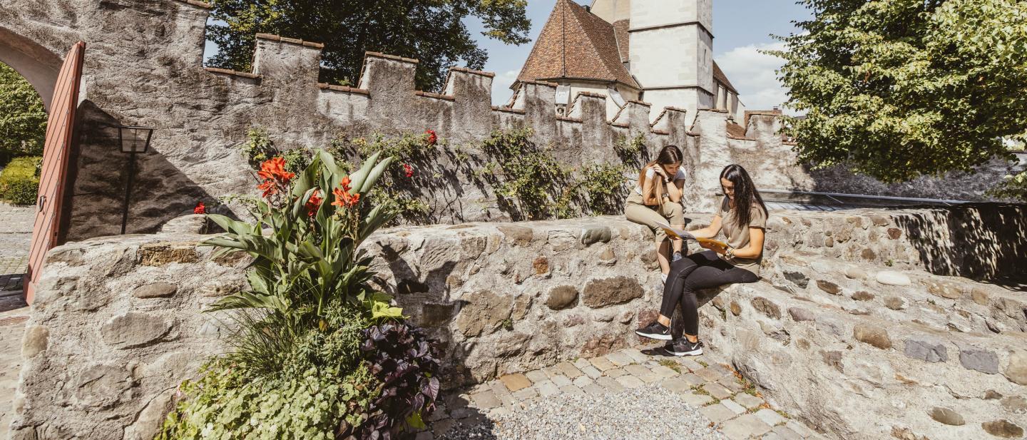 Zwei Frauen sitzen auf einer Steinmauer und studieren eine Broschüre. Im Hintergrund ist eine Kirche zu sehen.