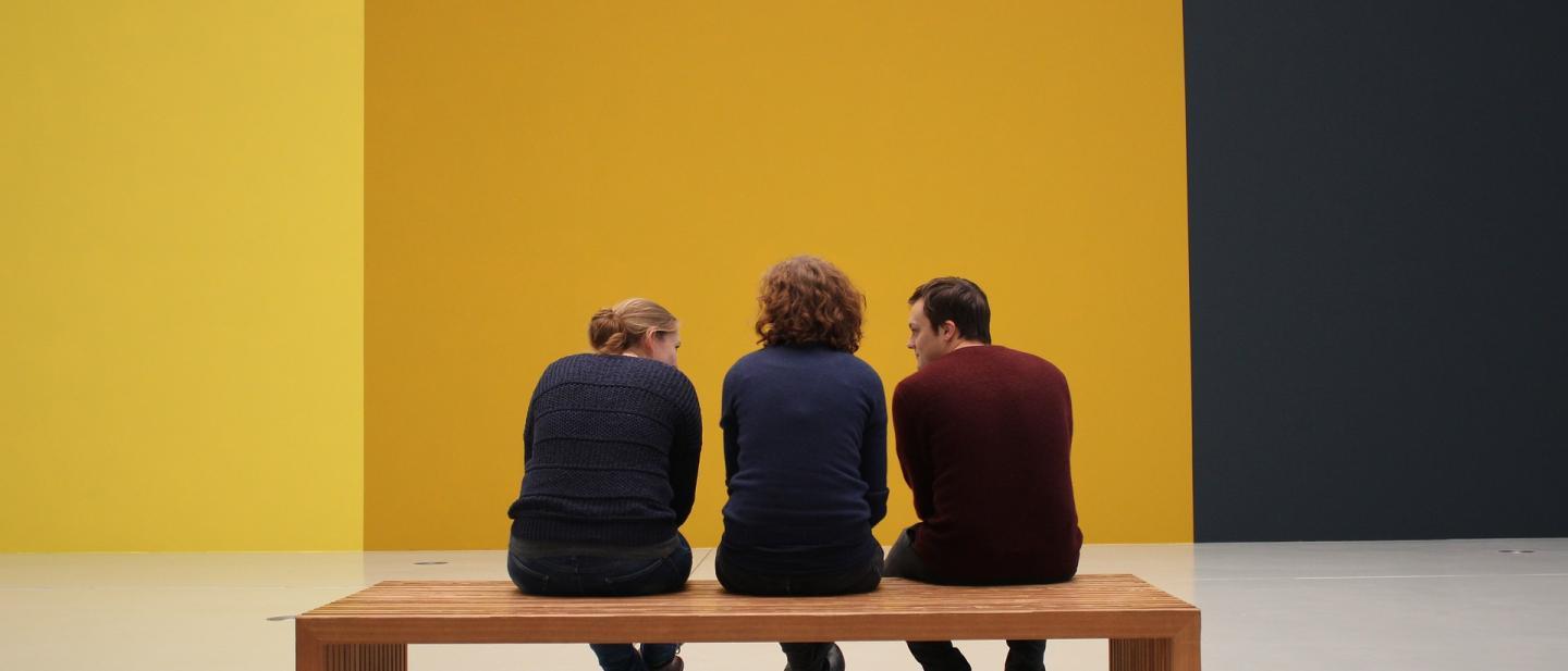 Drei Menschen sitzen auf einer Holzbank im Museum und betrachten eine in Gelbfarben abgestufte Wand.