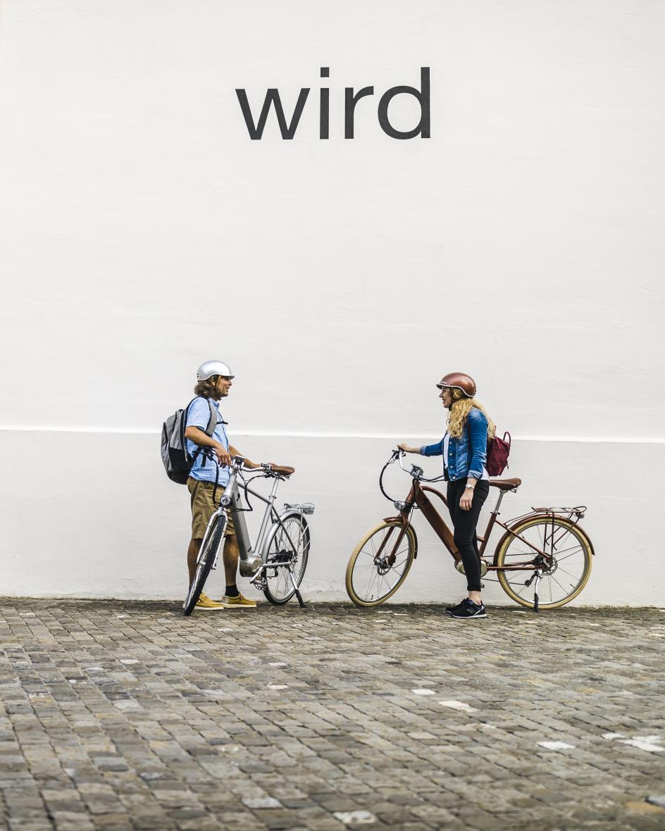 Zwei Radfahrende stehen vor einer wissen Wand mit dem Schriftzug wird des Kunsthauses Zug.
