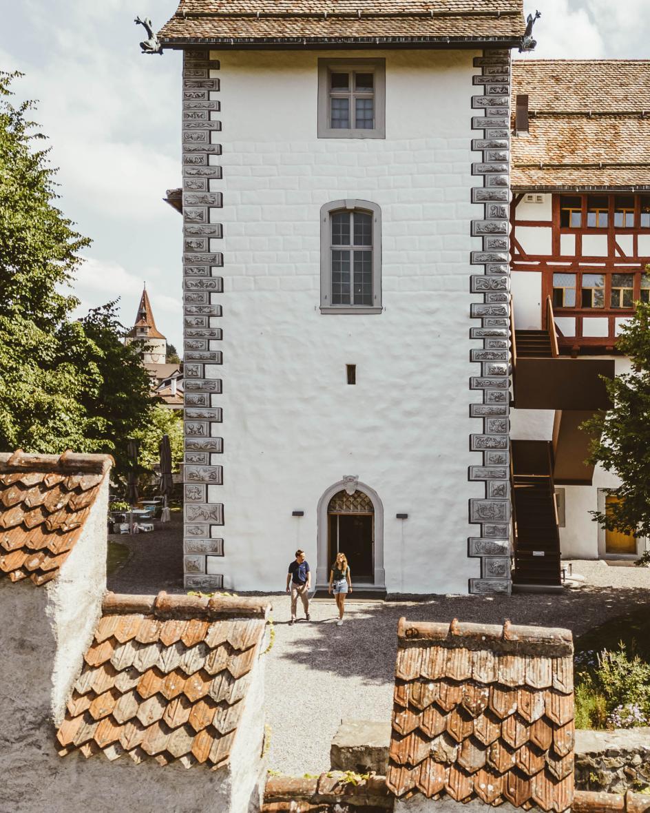 Ein Mann und eine Frau laufen aus dem historischen Steinturm des Museum Burg Zug auf dem Kiesweg Richtung Torbogen.