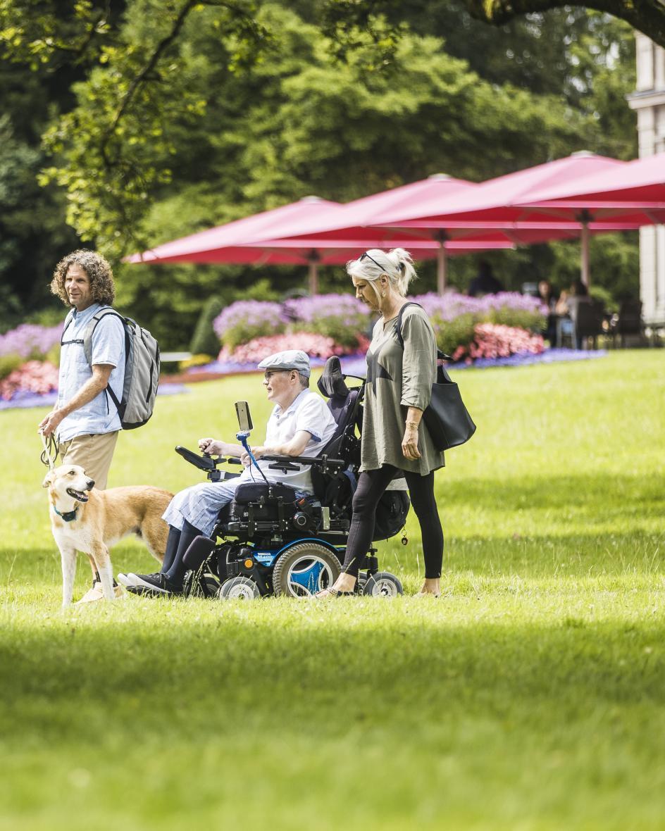 Drei Menschen mit Hund spazieren durch den grünen Villette Park in Cham. Ein Mann sitzt im Rollstuhl.